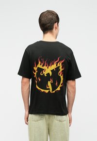 Μαύρο βαμβακερό t-shirt με μεγάλο, πολύχρωμο σχέδιο φλόγας και κίτρινο και κόκκινο λογότυπο στην πλάτη. Κοντά μανίκια και άνετη εφαρμογή.