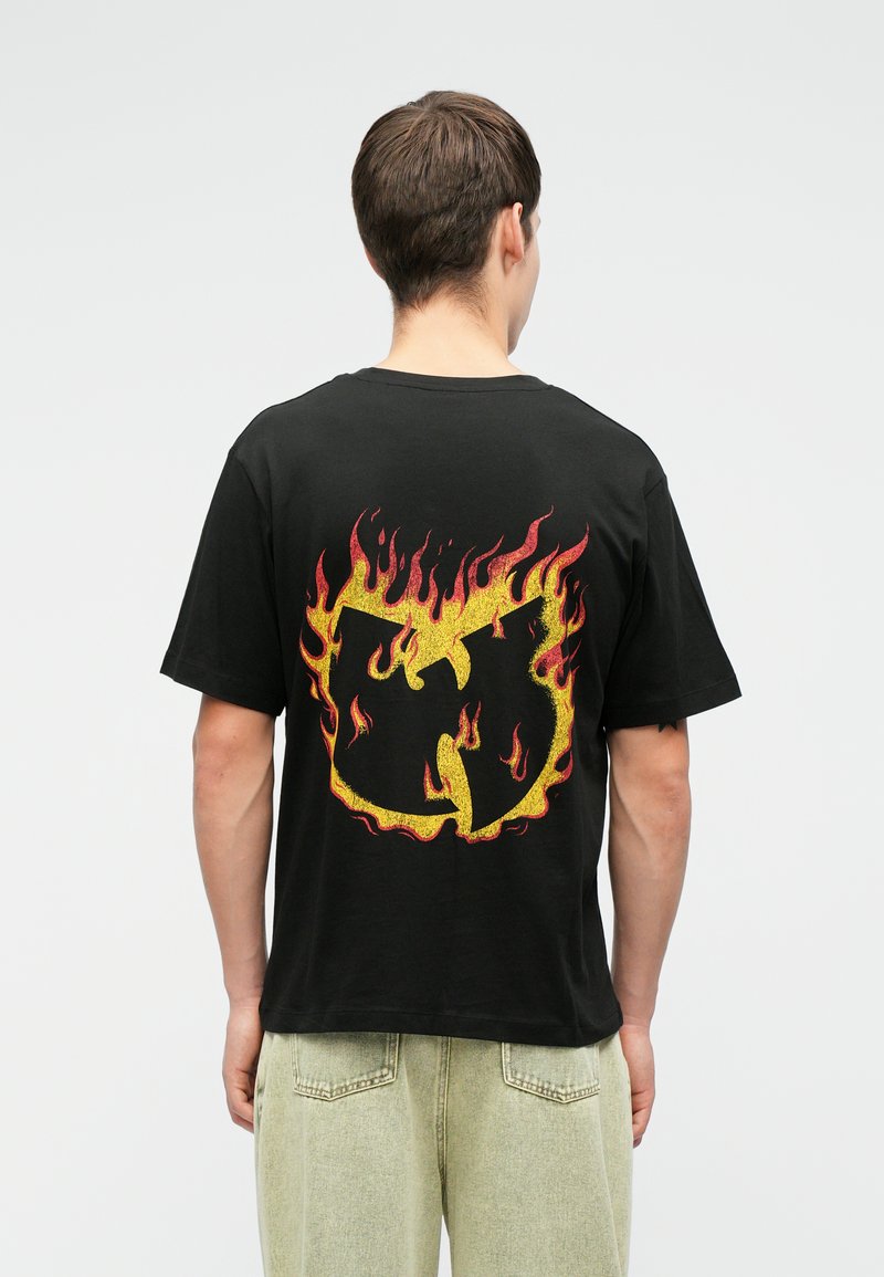 Μαύρο βαμβακερό t-shirt με μεγάλο, πολύχρωμο σχέδιο φλόγας και κίτρινο και κόκκινο λογότυπο στην πλάτη. Κοντά μανίκια και άνετη εφαρμογή.