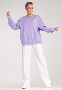 Lavenderowa bluza w luźnym kroju z ribbowymi mankietami i dolnym brzegiem, połączona z białymi spodniami o wysokim stanie i białymi sneakersami.
