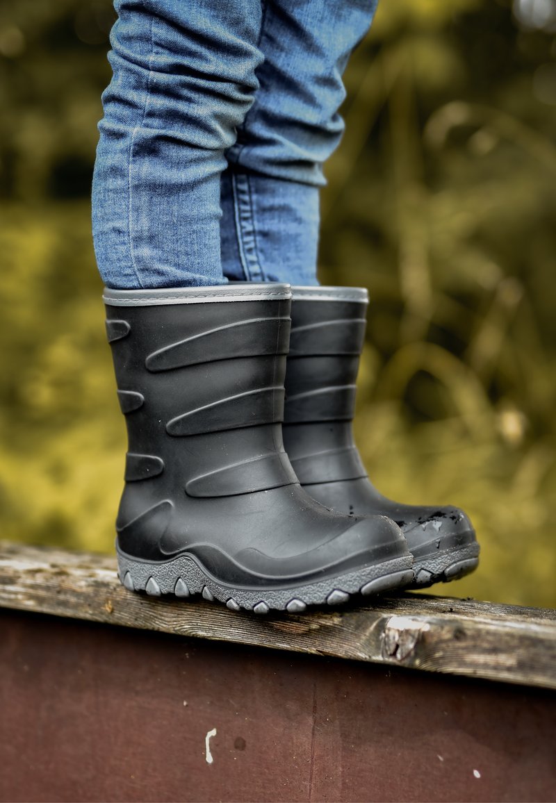 Mustat kumisaappaat, joissa on kuvioitu pinta, harmaat pohjat ja uritettu muotoilu. Asetettu puupinnalle, osittain sinisten farkkujen peitossa.