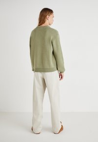 Pulover verde tricotat cu guler rotund, umeri lăsați și tiv cu nervuri, asortat cu pantaloni largi crem și pantofi maro.