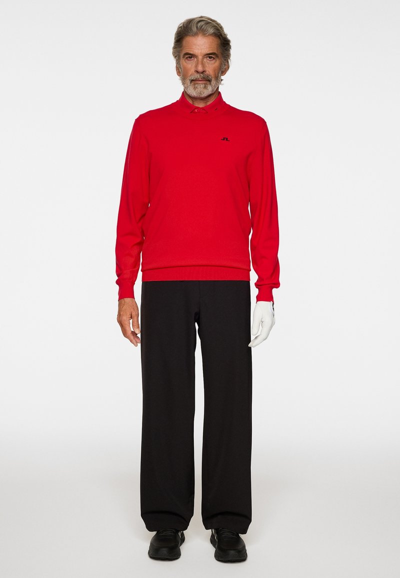 Mann steht da und trägt ein rotes langärmliges Hemd, schwarze weite Hosen, schwarze Schuhe und einen weißen Handschuh an der rechten Hand vor einem schlichten Hintergrund.