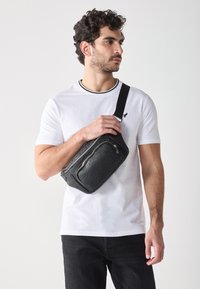Sac bandoulière noir compact en matériau texturé, avec des fermetures éclair, une sangle réglable et un motif en relief.