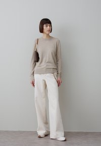 Donna con capelli corti e scuri che indossa un maglione beige a maniche lunghe, pantaloni bianchi a gamba larga, scarpe bianche e porta una piccola borsa a tracolla marrone.