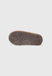 Semelle en caoutchouc brun avec des nervures texturées, arborant le logo "UGG". Conception large pour la stabilité. Motif de semelle notable qui améliore l'adhérence.