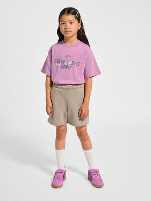 Ung pige står iført en lilla "Serve the Game" t-shirt, beige shorts, hvide sokker, lilla sneakers og lyserøde hårclips.