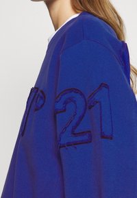 Mujer vistiendo un sudadero azul con tejido en relieve y el número "21" y letras visibles en la parte delantera y la manga.