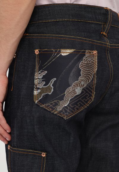 Jeans en denim foncé avec des coutures orange contrastantes. La poche notable présente un design graphique métallique complexe et un bord à motifs.