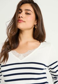 Top de manga larga a rayas en blanco y azul marino, con escote en V y bordes de encaje, y un diseño ajustado. Tela suave con una textura lisa.