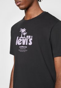 Jeune personne portant un t-shirt noir Levi's avec un logo de palmier rose et le texte « California International » sur la poitrine.