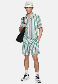 Gestreepte korte mouwen shirt en shorts in groen, wit en paars; gecombineerd met een crème kleurige bucket hat en een zwarte tote bag. Sneakers met logo sokken.
