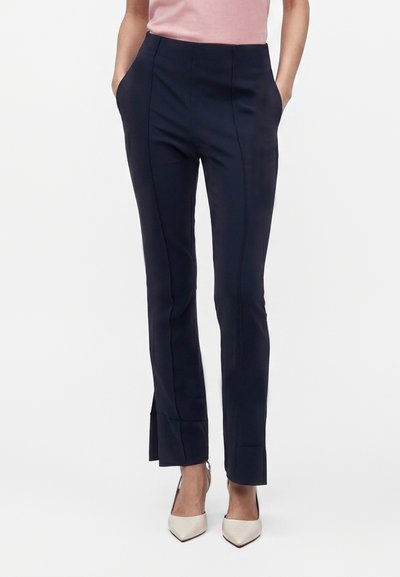Femme portant un pantalon noir tailleur avec coutures devant et fentes sur les côtés, associé à des talons blancs à bout pointu et un haut rose.