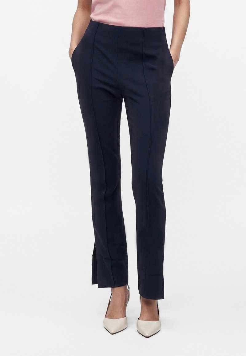 Femme portant un pantalon noir tailleur avec coutures devant et fentes sur les côtés, associé à des talons blancs à bout pointu et un haut rose.