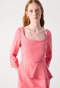 Robe rose avec un décolleté carré et des manches trois-quarts. Présente un détail de peplum superposé à la taille, offrant une silhouette structurée.