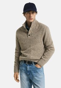 Mann in einem beigen Strickpullover mit Knöpfen, blauen Jeans, schwarzem Gürtel, marineblauem Hut und einer Hand in der Tasche vor einem schlichten Hintergrund.