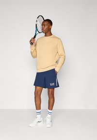 Beige genser og marineblå shorts med EA7-logo, hvite stripe detaljer, kombinert med hvite treningssko og blåstripete sokker.