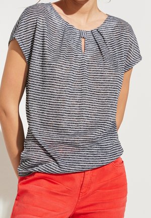 Femme portant un haut à manches courtes rayé noir et blanc avec un décolleté en trou de serrure, associé à un pantalon rouge vif.