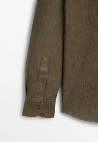 Manche longue d'une chemise en tissu brun texturé avec poignet boutonné et patte de manche, présentée sur fond blanc.