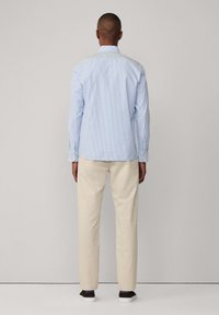 Camicia azzurra a strisce, con bottoni e maniche lunghe, abbinata a pantaloni beige e calzature scure, vista da dietro.