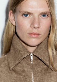 Giacca in maglia marrone con colletto alto e zip argentata. Tono della pelle chiaro, occhi blu e capelli biondi lisci incorniciano il viso.