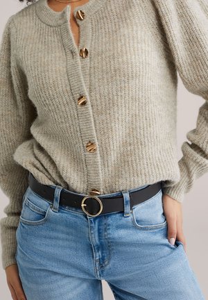 Žena nosící béžový pletený cardigan se zlatými knoflíky, zastrčený do světle modrých džín s černým pásem se zlatou kulatou přezkou.