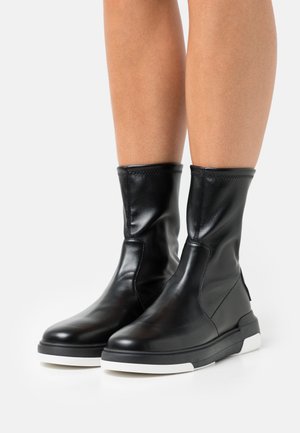 Stivaletti neri in pelle fino a metà polpaccio con suole spesse in gomma nera e bianca, indossati su gambe nude su sfondo bianco.