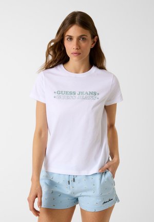 Frau trägt ein weißes "Guess Jeans"-T-Shirt und hellblaue Shorts mit floralem Muster, steht mit einer Hand in der Tasche.