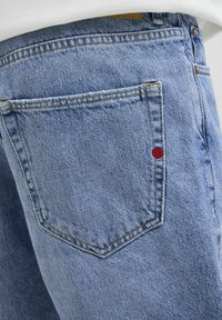 Jeans de mezclilla azul claro con un bolsillo trasero que presenta costuras amarillas en contraste y un pequeño acento rojo cerca de la costura del bolsillo.