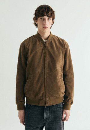 Joven con cabello castaño ondulado, vistiendo una chaqueta bomber de ante marrón y jeans oscuros, posando frente a un fondo claro y liso.