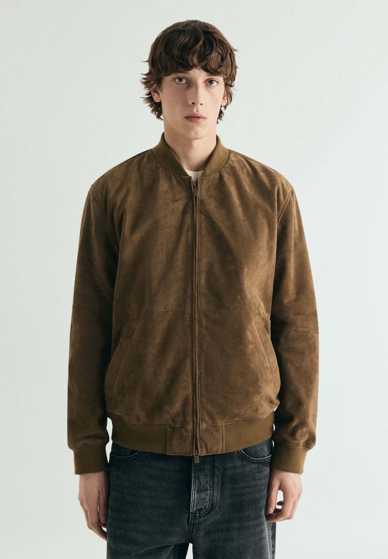 Jonge man met golvend bruin haar, gekleed in een bruine suède bomberjack en donkere spijkerbroek, tegen een effen lichte achtergrond.