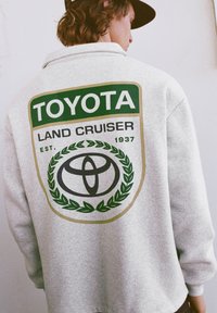 Felpa grigia con un grande logo Toyota Land Cruiser verde, oro e nero e motivo di alloro sul retro. Sempile design con collo tondo.