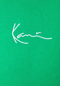 Tejido verde con una textura suave y tersa que presenta una firma bordada en blanco de "Kamis". El fondo es uniforme y sin patrones.