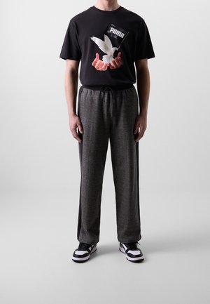 Mann trägt schwarzes Puma T-Shirt mit Händen, die eine weiße Tauben-Grafik halten, graue Jogginghose mit Seitenstreifen und schwarz-weiße Sneakers.