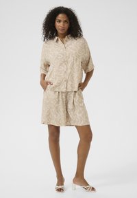 Chemise à manches courtes boutonnée à motifs beiges et shorts assortis, tous deux ornés de motifs tourbillonnants blancs. Le modèle se tient les mains dans les poches.