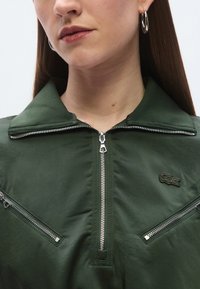 Πράσινο μπουφάν με φερμουάρ και ψηλό γιακά, με μεταλλικά φερ μουάρ και μικρό κεντημένο λογότυπο. Ομαλή υφή και κομψός σχεδιασμός.