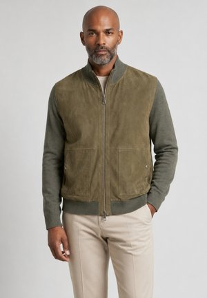Mann mit kurzem Bart, der eine olivgrüne Jacke mit Reißverschluss und beige Hose trägt, steht mit einer Hand in der Tasche vor einfarbigem Hintergrund.