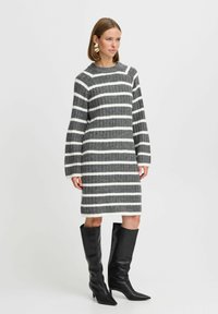 Strickkleid mit grau-weißen Streifen, langen Ärmel, gerippter Textur und rundem Ausschnitt. Kombiniert mit kniehohen schwarzen Stöckelschuhen.