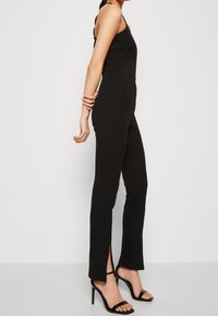 Femme portant un haut noir sans manches et un pantalon noir à fentes latérales, assortis à des sandales à talons hauts noires, debout de profil.