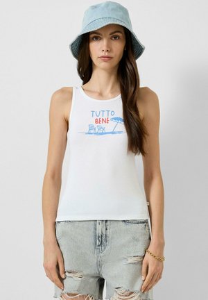 Femme portant un chapeau bob bleu clair, un débardeur blanc avec l'inscription "TUTTO BENE" et un motif de chaise de plage, ainsi que des shorts en jean délavés et usés.