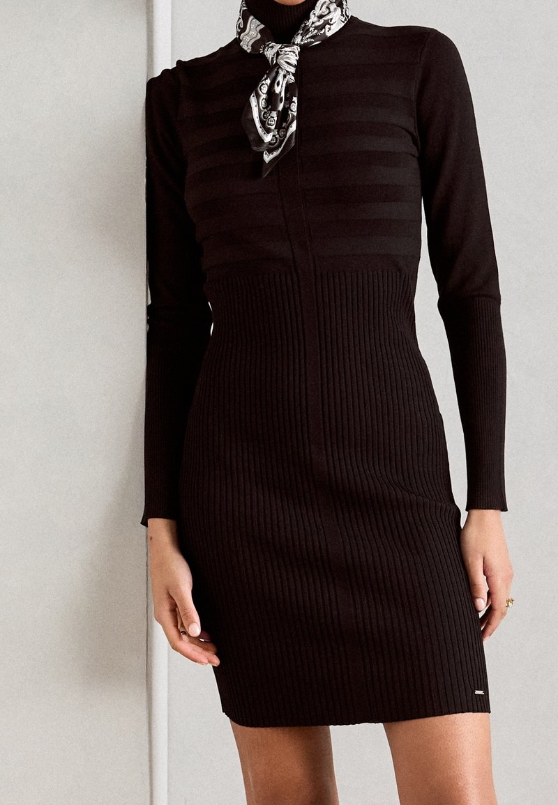 Femme portant une robe en maille côtelée noire à manches longues et une écharpe noire et blanche à motifs nouée autour du cou.