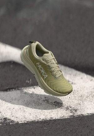 Hellgrüner Hoka-Laufschuh mit weißem Logo schwebt über einer schwarz-weißen Asphaltstraße.