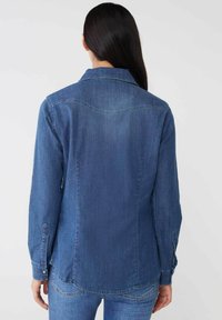 Camicia in denim di blu medio con colletto, maniche lunghe e polsini abbottonati. Presenta un orlo curvo e dettagli sul giogo posteriore.