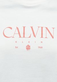 Hvit bomulls-T-skjorte med "CALVIN KLEIN" i korallrosa bokstaver, med "Est. 1968" og blomsterdetalj under. Enkel rund hals.