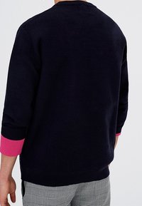 Persona con un maglione nero a maniche lunghe e polsini rosa, e pantaloni grigi a quadri, mostrata di spalle.