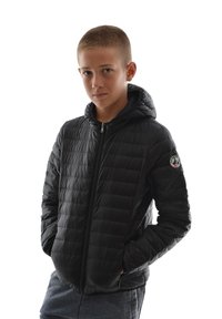 Doudoune noire avec une capuche, présentant une texture matelassée, une fermeture éclair à l'avant et un patch logo sur la manche gauche. Portée avec un pantalon de survêtement gris.