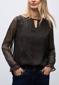 Blusa nera trasparente con stampa leopardata, scollatura a V con apertura a chiave e maniche lunghe volantee. Abbinata a jeans blu. Tessuto leggero e testurizzato.