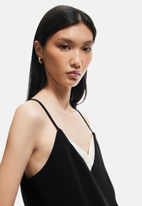 Top nero senza maniche con un inserto bianco a forma di V, dotato di spalline sottili e una texture liscia. Il modello mostra il capo di abbigliamento da un angolo laterale.