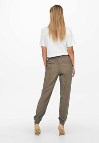 Femme aux longs cheveux blonds portant une chemise blanche à manches courtes, un pantalon jogger taille élastique vert olive et des talons hauts beige, vue de dos.