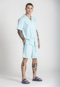 Camicia blu chiaro a maniche corte con colletto e bottoni, abbinata a pantaloni corti coordinati. Indossata con sandali blu a scivolo. Tatuaggi visibili sulle gambe.