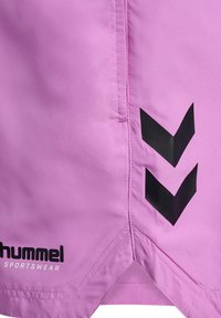 Rosa sportshorts i mjuk tyg, med svarta chevrondetaljer och logotext, samt en triangulär skärning vid fållen för extra detalj.
